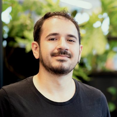 Mert Erbil, smiling, in a black t-shirt in front of green plants