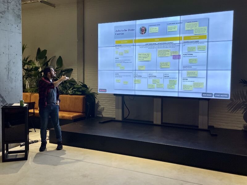 Mert on a stage pointing at a large screen showing a Jobs to be Done canvas filled with sticky notes.
