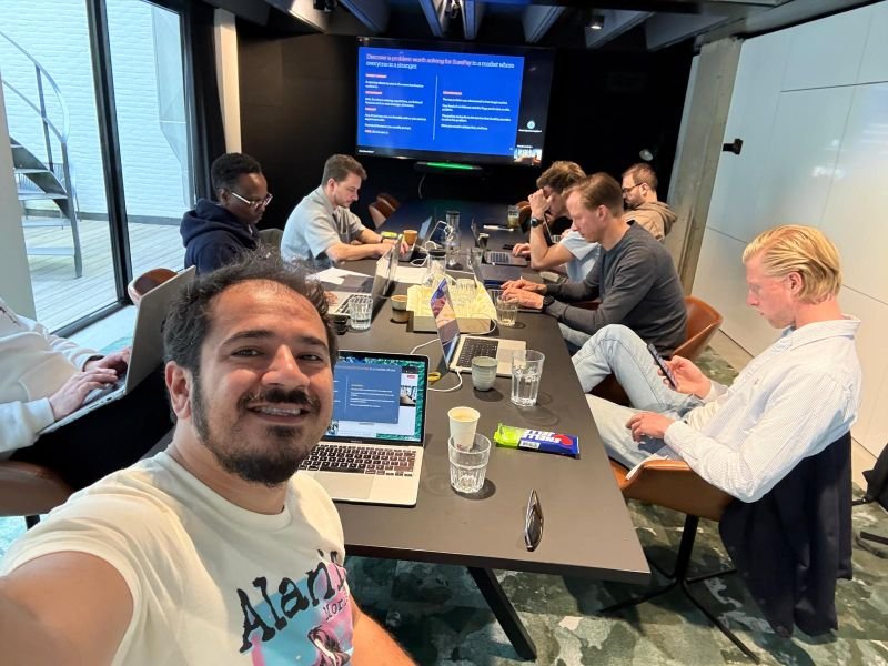 A selfie of Mert in front of a long meeting table where the SurePay product team works on laptops, with workshop slides on the screen behind them.