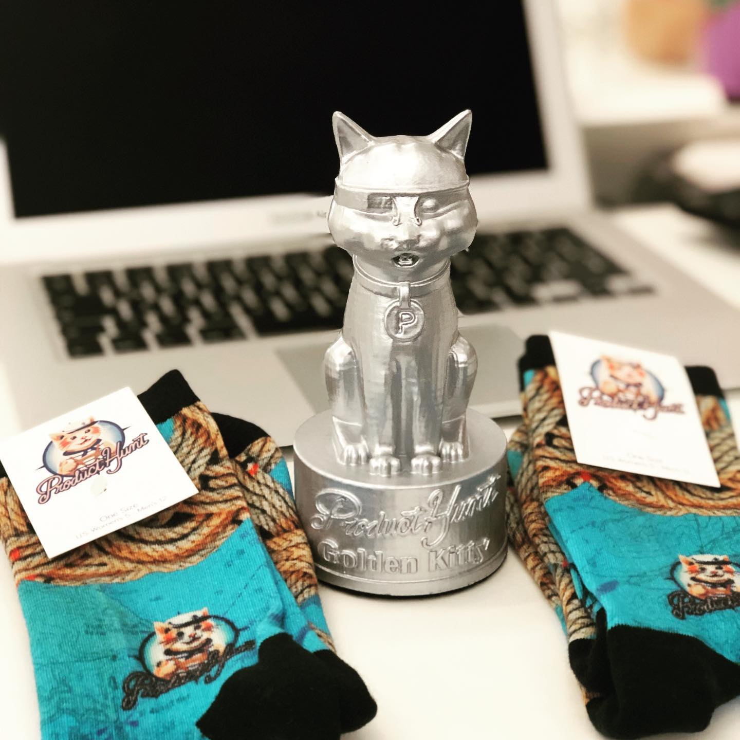 The silver Product Hunt Golden Kitty statue on a desk in front of a laptop, between two pairs of Product Hunt cat socks
